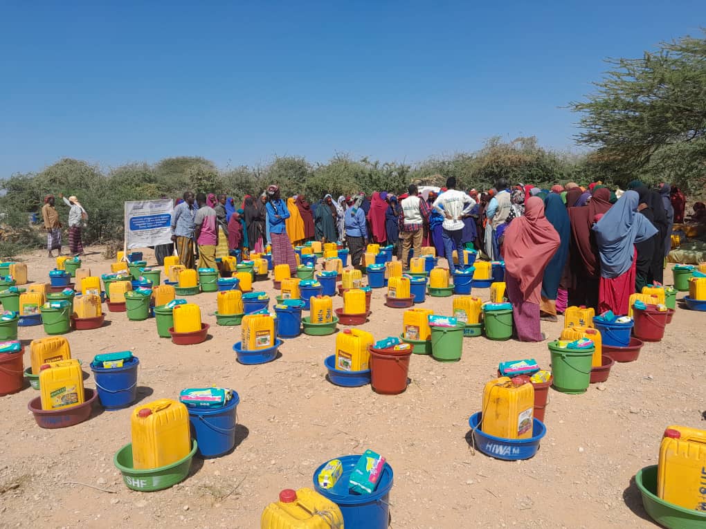 Community members beside water containers and household supplies arranged for distribution
